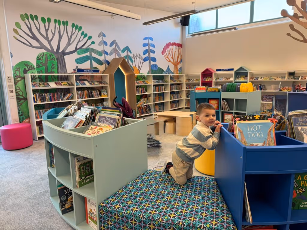 Child kneeling on patterned carpet in a bright, colorful children's library with books and murals.