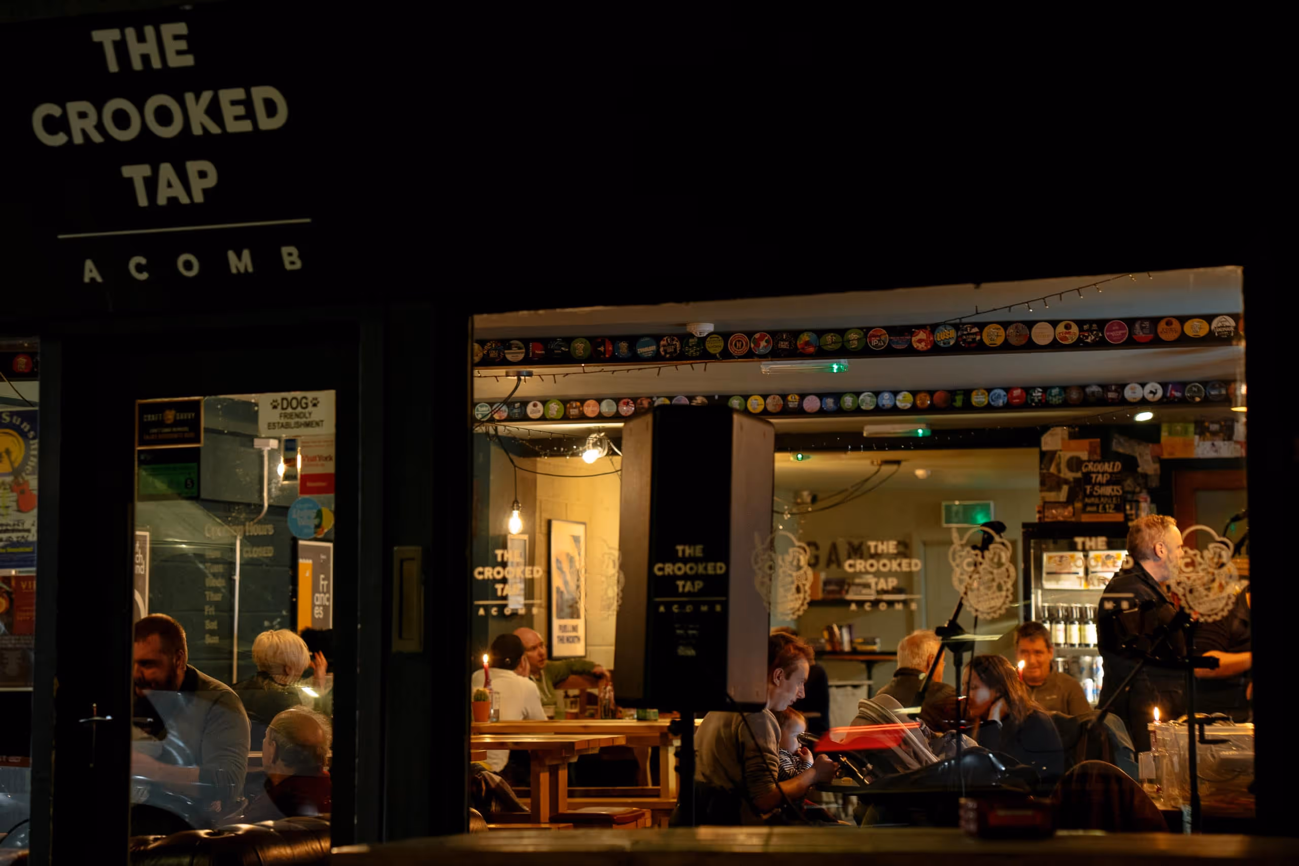 Night view through window of The Crooked Tap pub with seated patrons and warm interior lighting.