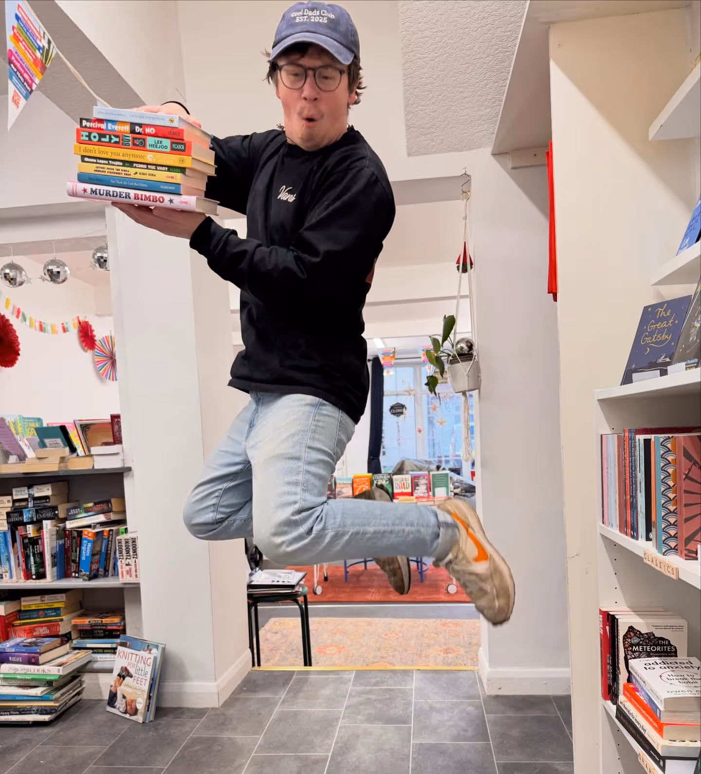 Man wearing glasses and cap jumps inside a bookstore holding a stack of colorful books.