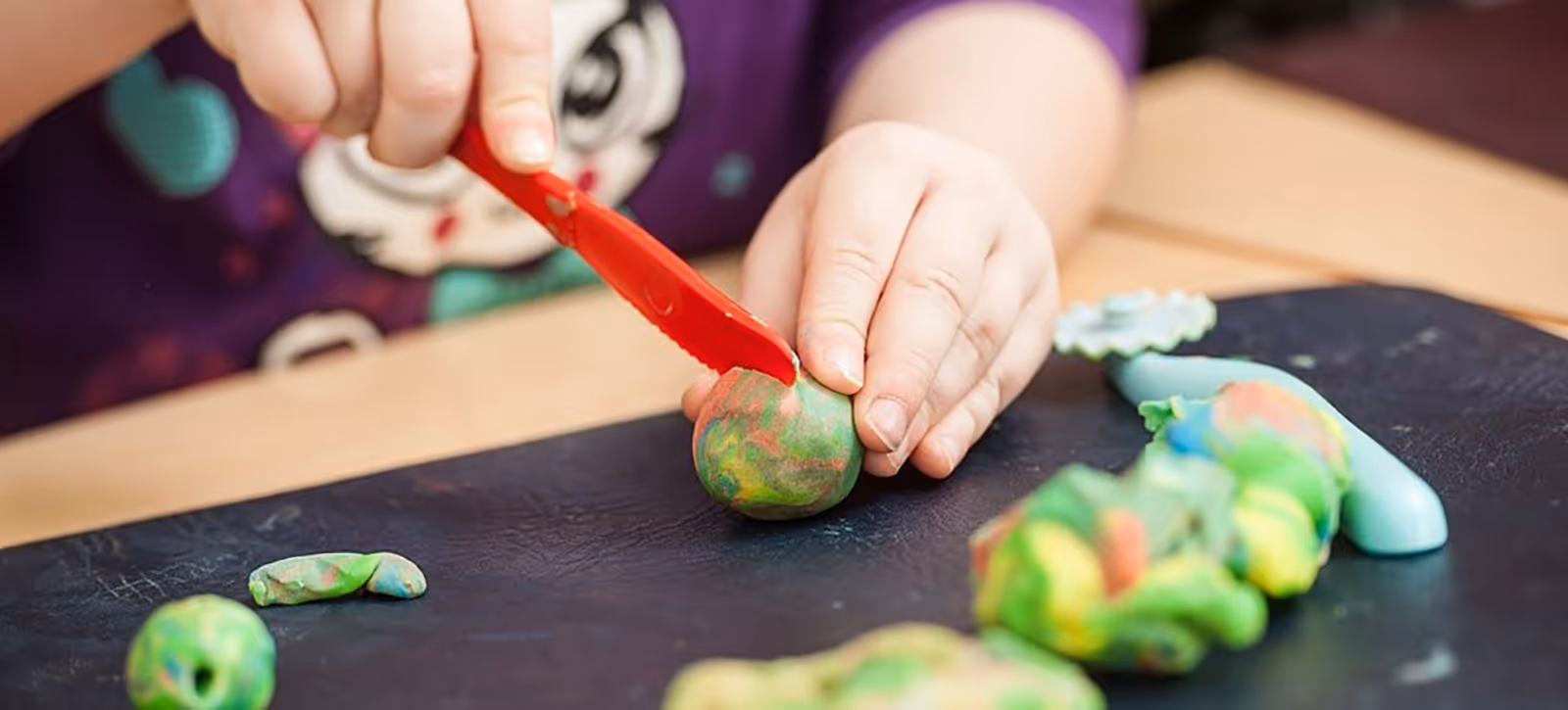 Child shaping colorful clay with a red plastic knife on a black surface.