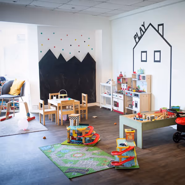 Bright children's playroom with wooden toys, small tables, and a wall drawing of a house labeled 'PLAY'.