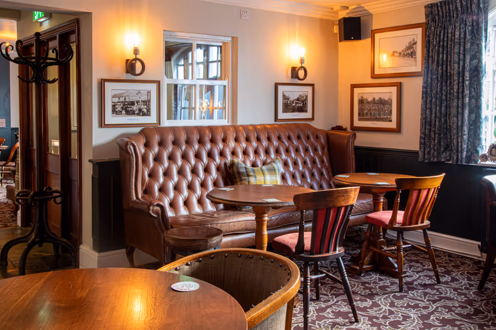 Cozy pub interior with tufted leather sofa, wooden tables, chairs, framed photos, and patterned carpet.