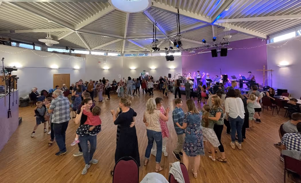 Group of people dancing and socializing in a large hall with a live band playing on stage.