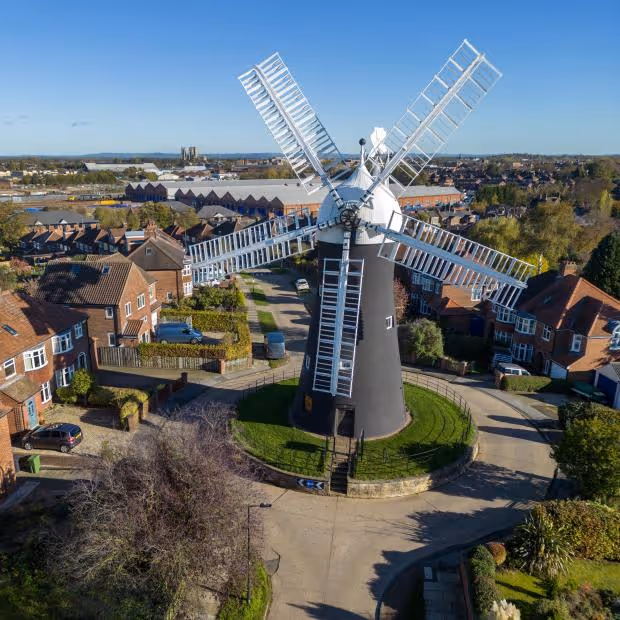 Historic black windmill with white sails in suburban neighborhood under clear blue sky.