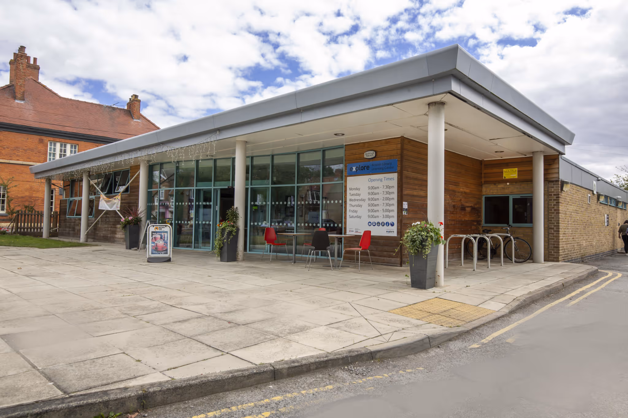 Modern community library building with glass entrance, outdoor seating, planters, and opening times sign.