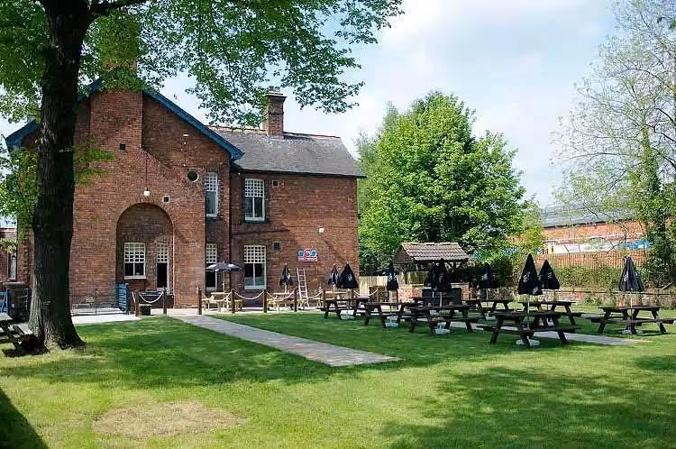 Outdoor seating area with wooden picnic tables and umbrellas beside a red brick building and green trees.