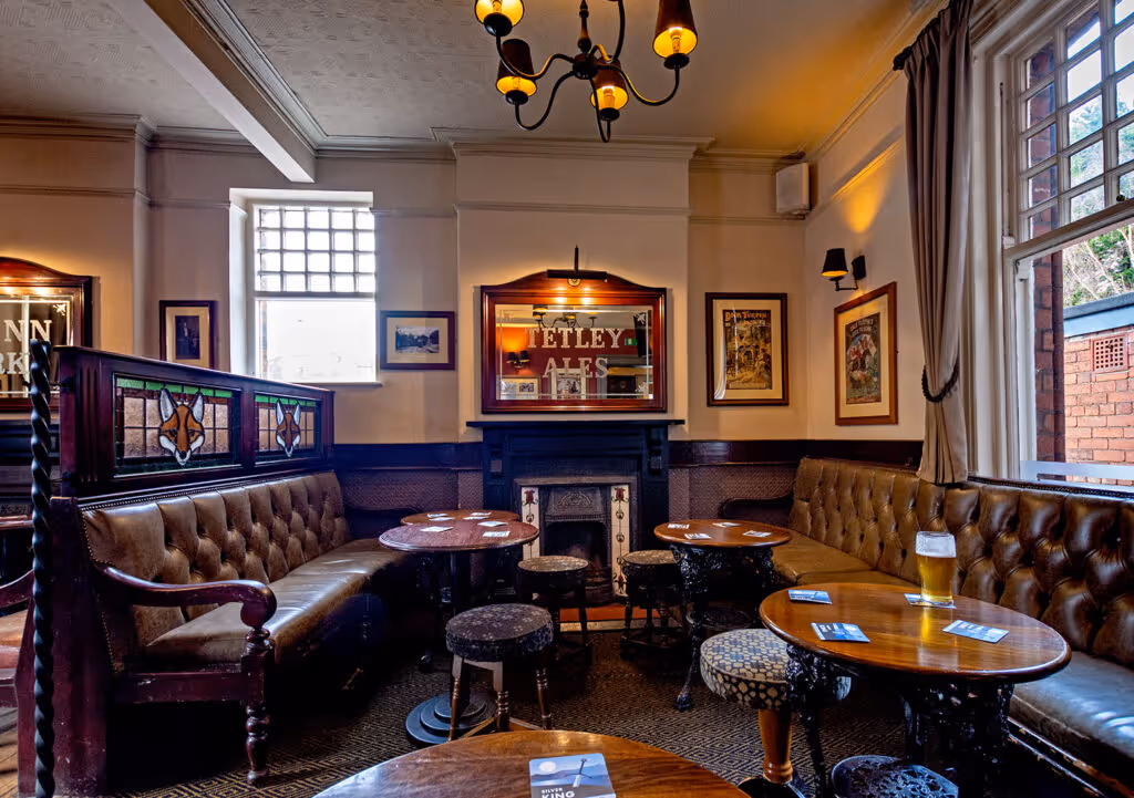 Cozy pub interior with brown leather seating, wooden tables, stained glass fox decorations, and a pint of beer.