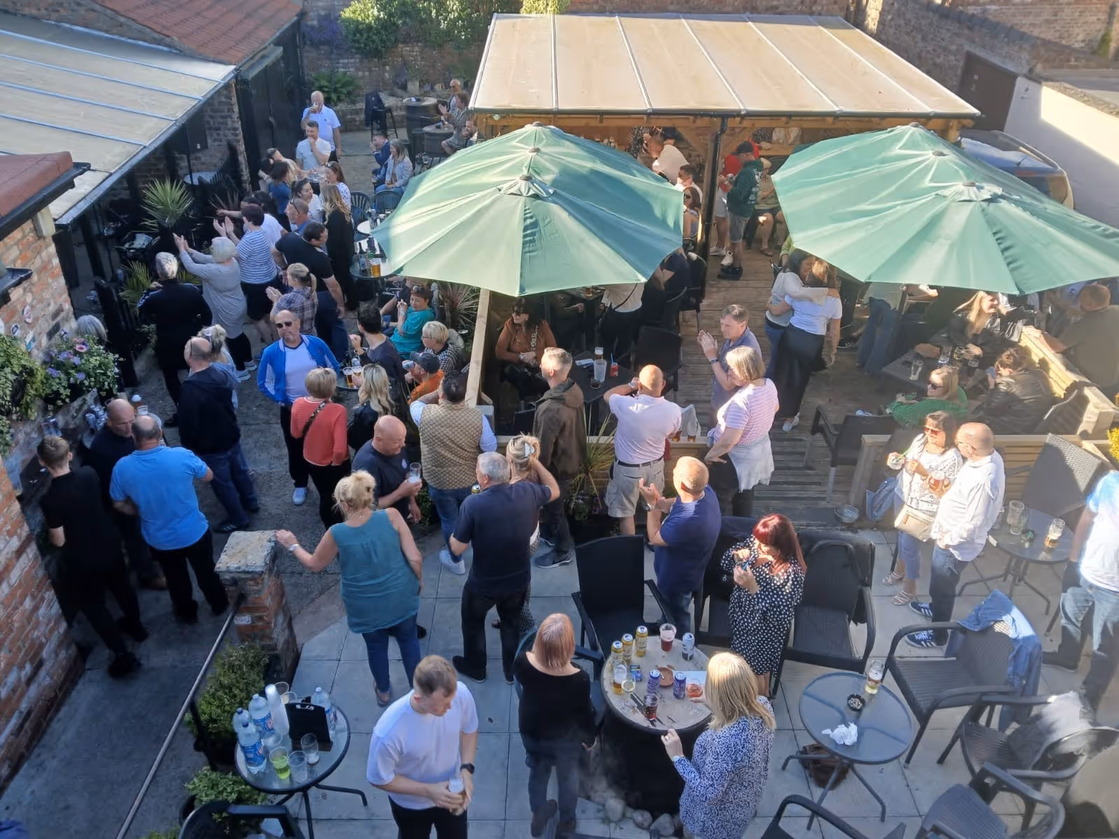 Crowded outdoor patio with people socializing under large green umbrellas on a sunny day.