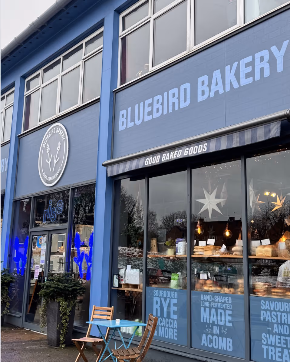 Bluebird Bakery storefront with large windows showing breads and a small outdoor table with two chairs.