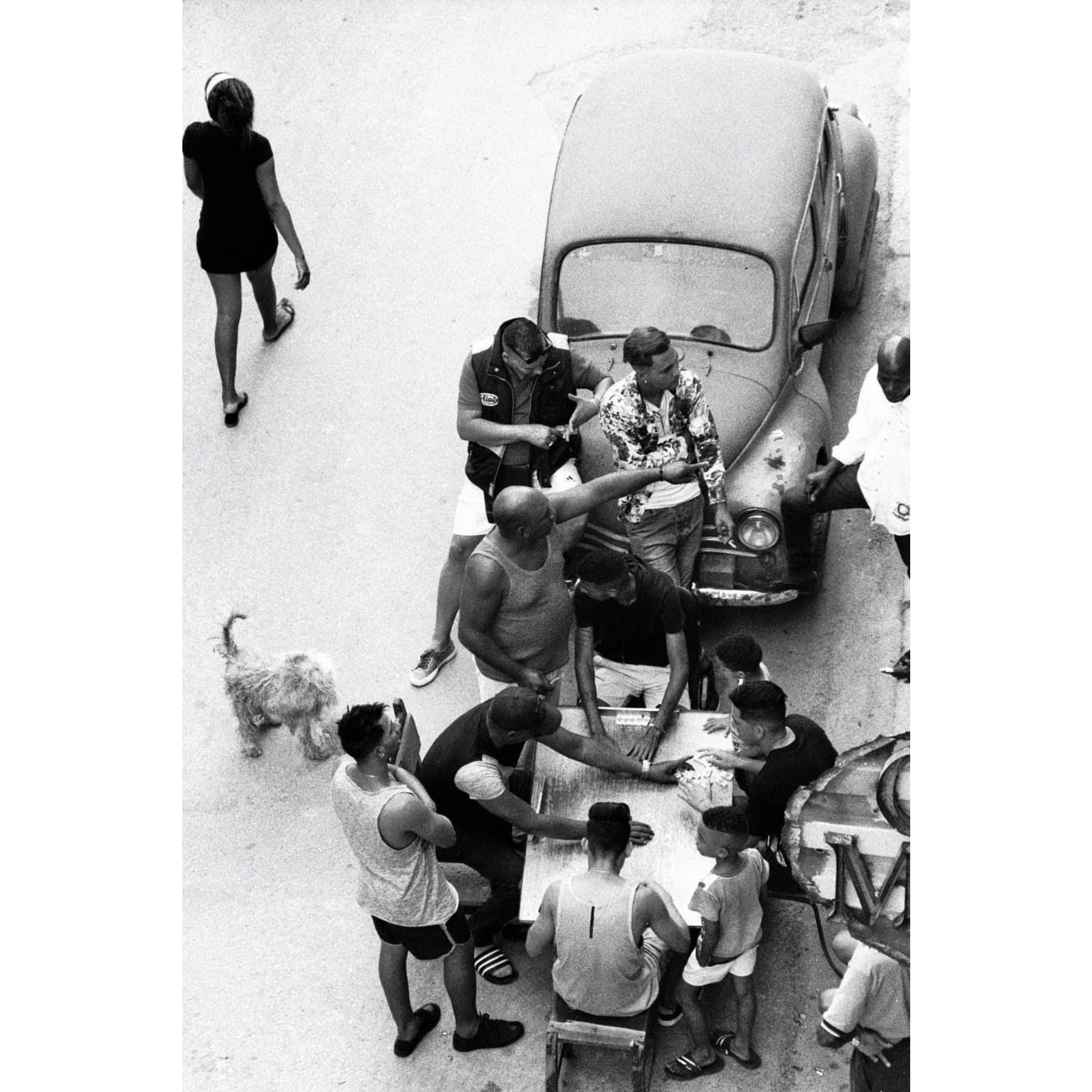 Black and white photo of people playing dominoes around a table near a vintage car outdoors.
