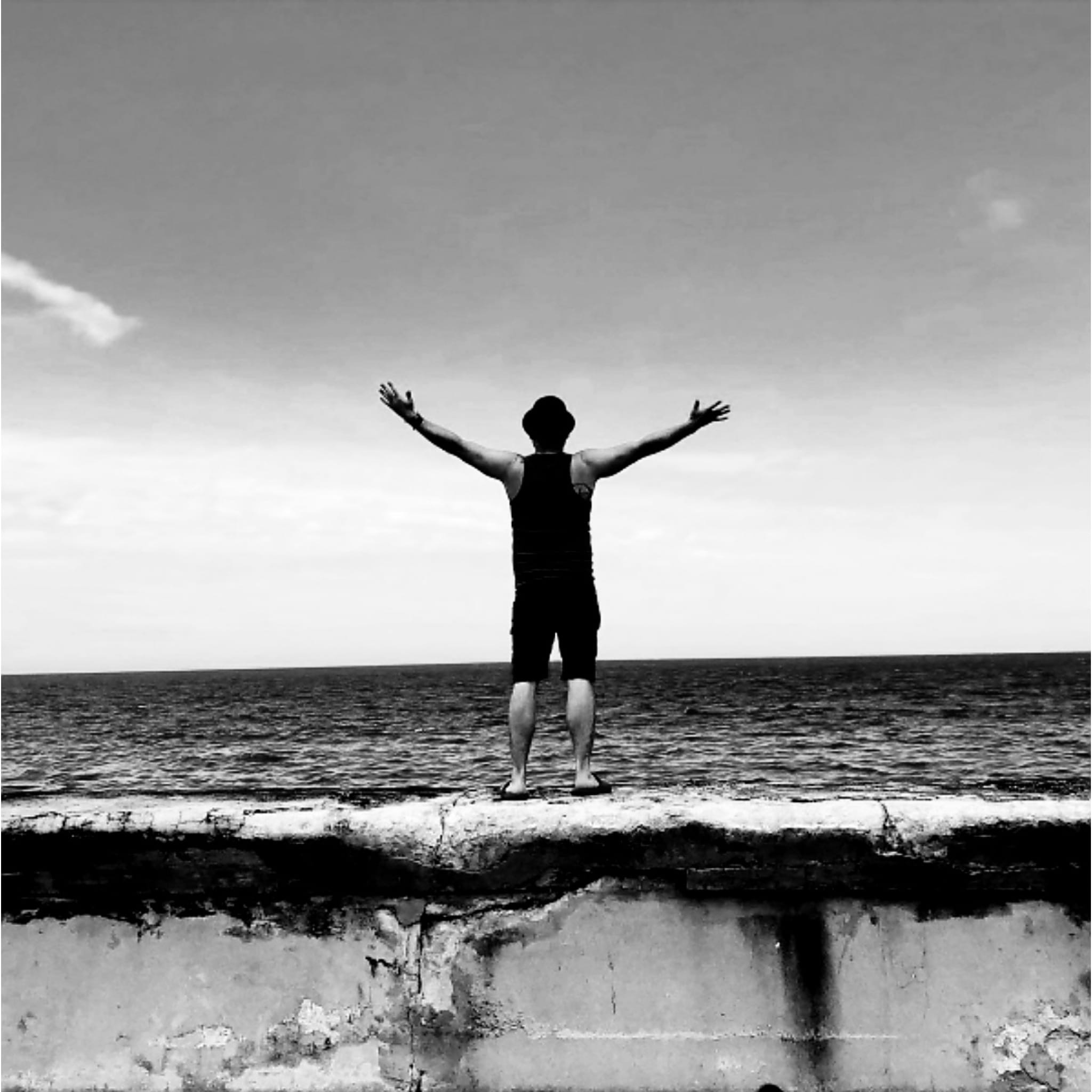Man with arms outstretched facing the ocean while standing on a stone wall under the sky.