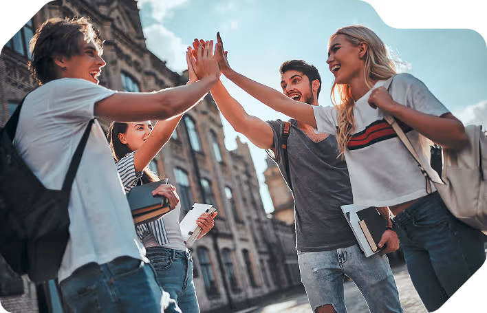 Grupa uśmiechniętych studentów z książkami przybija piątkę na tle miejskich kamienic.