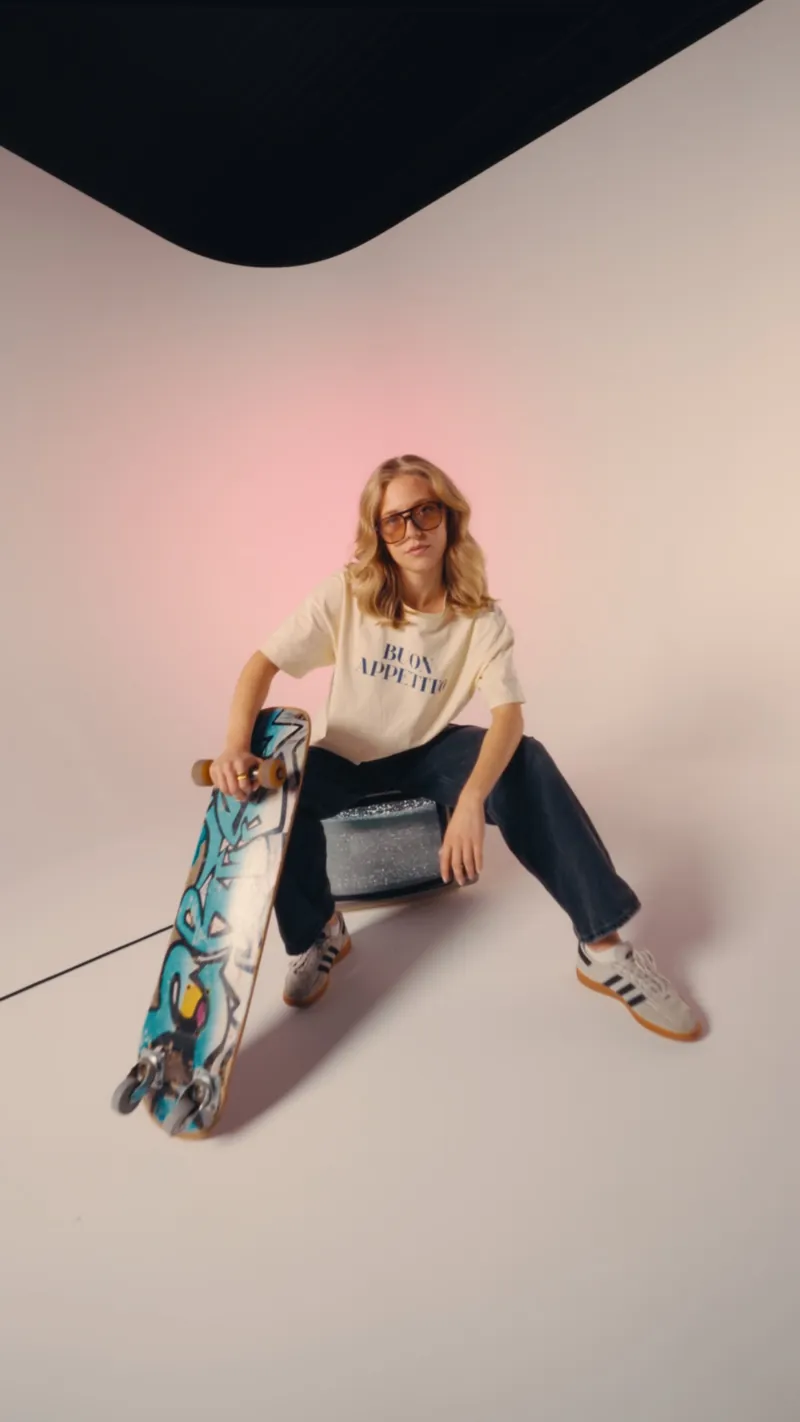 Model mit Skateboard sitzt auf einem Röhrenfernseher vor rosa beleuchtetem Hintergrund