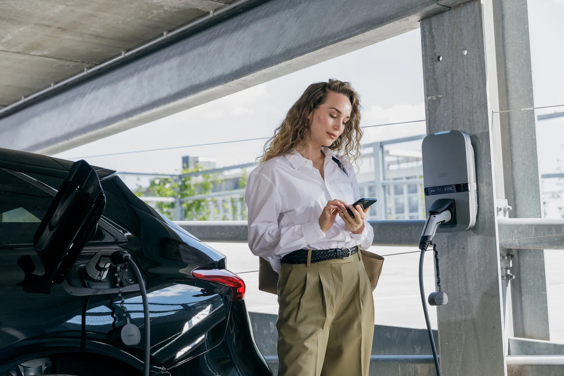Vrouw bij elektrische auto die oplaadt met een laadstation terwijl ze op haar telefoon kijkt.