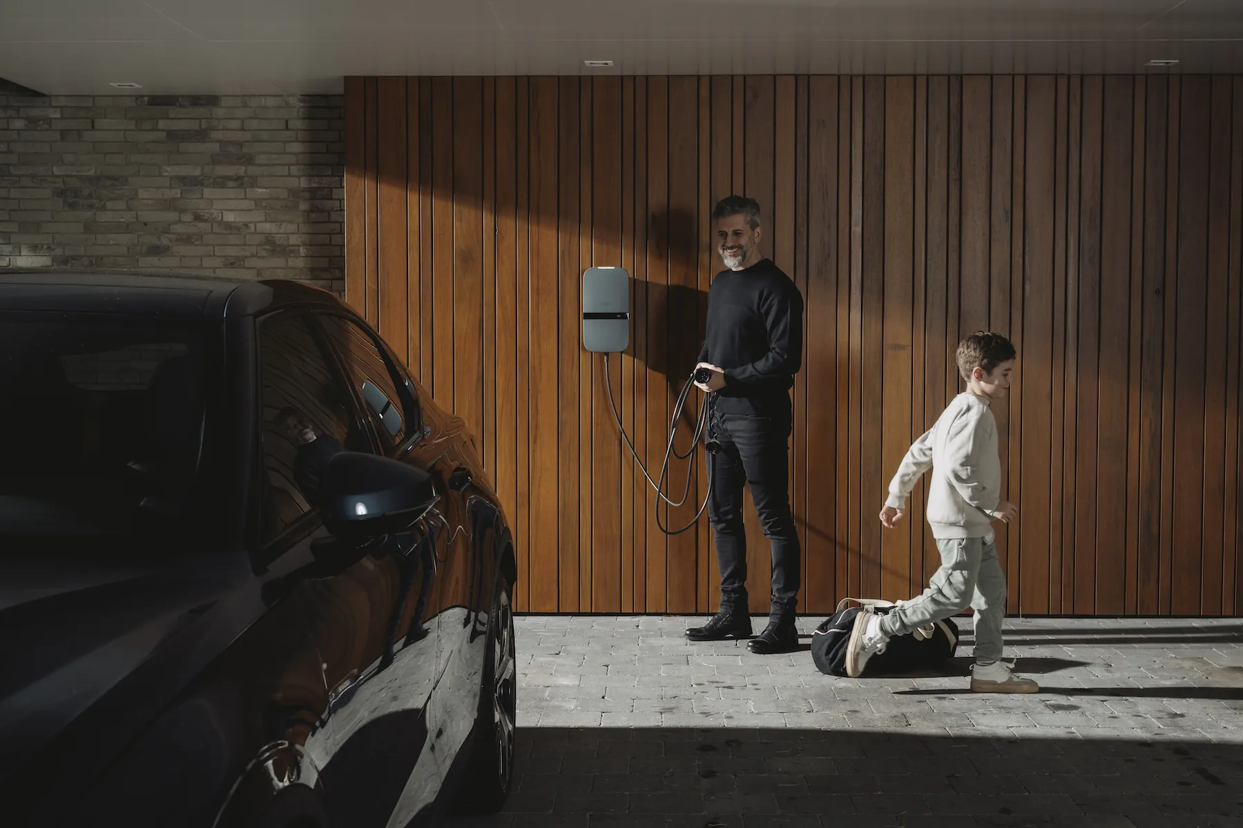 Man laadt elektrische auto op bij thuislader terwijl jongen langsloopt op oprit met houten muur.