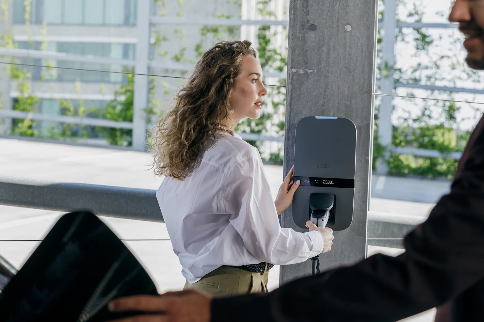 Vrouw laadt elektrische auto op bij een moderne laadpaal buiten met groen op de achtergrond.