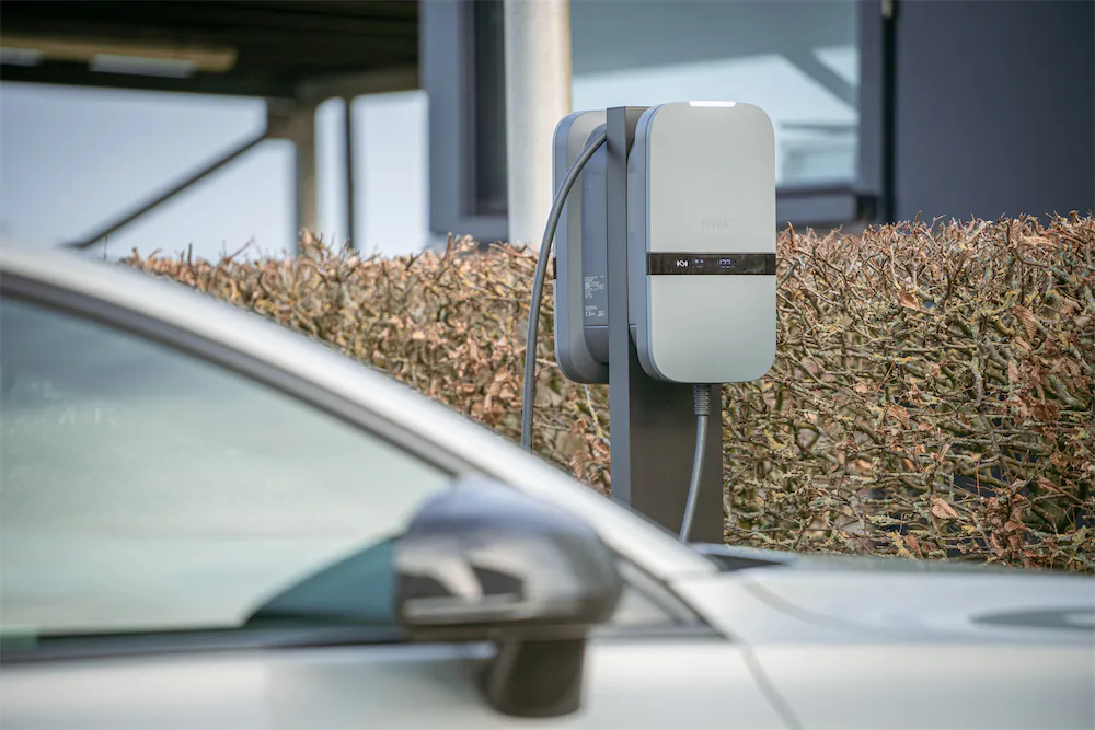 Elektrische auto verbonden met een grijze laadpaal naast een heg bij een modern gebouw.