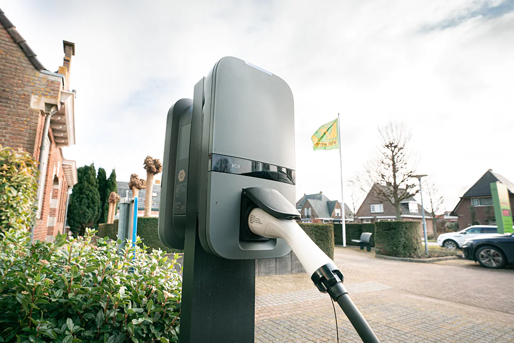 Elektrische auto-oplader paaltje met stekker in woonwijk met huizen en geparkeerde auto's.