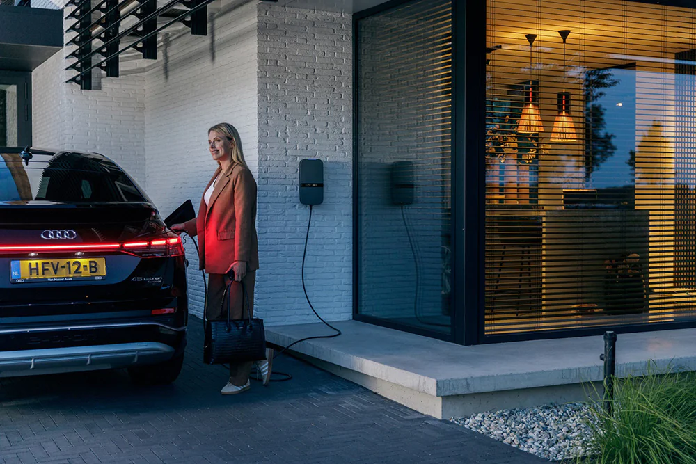 Vrouw laadt een zwarte Audi elektrische auto op naast een modern huis tijdens de avond.