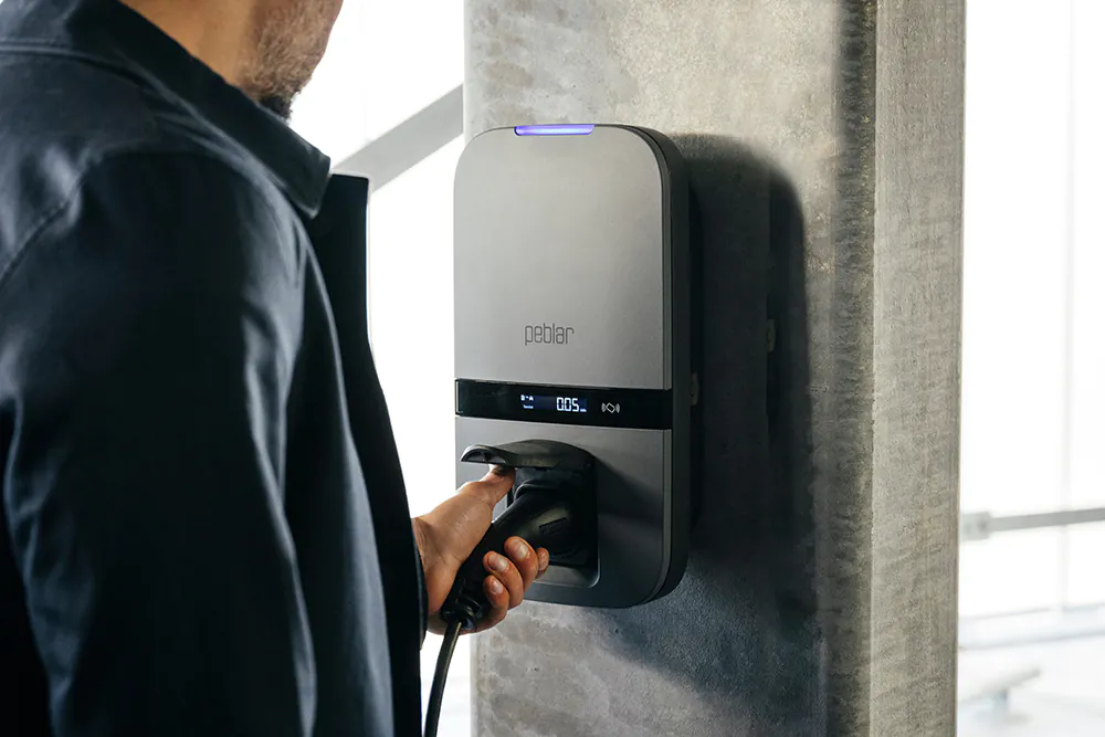 Persoon steekt een elektrische auto-oplaadstekker in een grijs oplaadstation aan een betonnen muur.