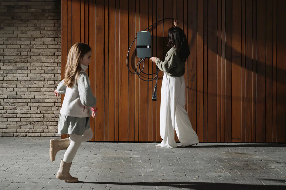 Vrouw hangt een oplaadkabel op en een meisje rent op een stenen pad voor een houten deur.