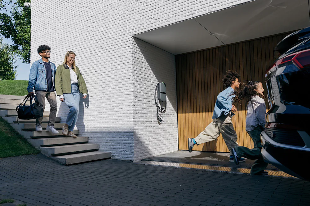 Twee kinderen rennen naar een elektrische auto bij een houten garage, terwijl twee volwassenen van de trap komen.