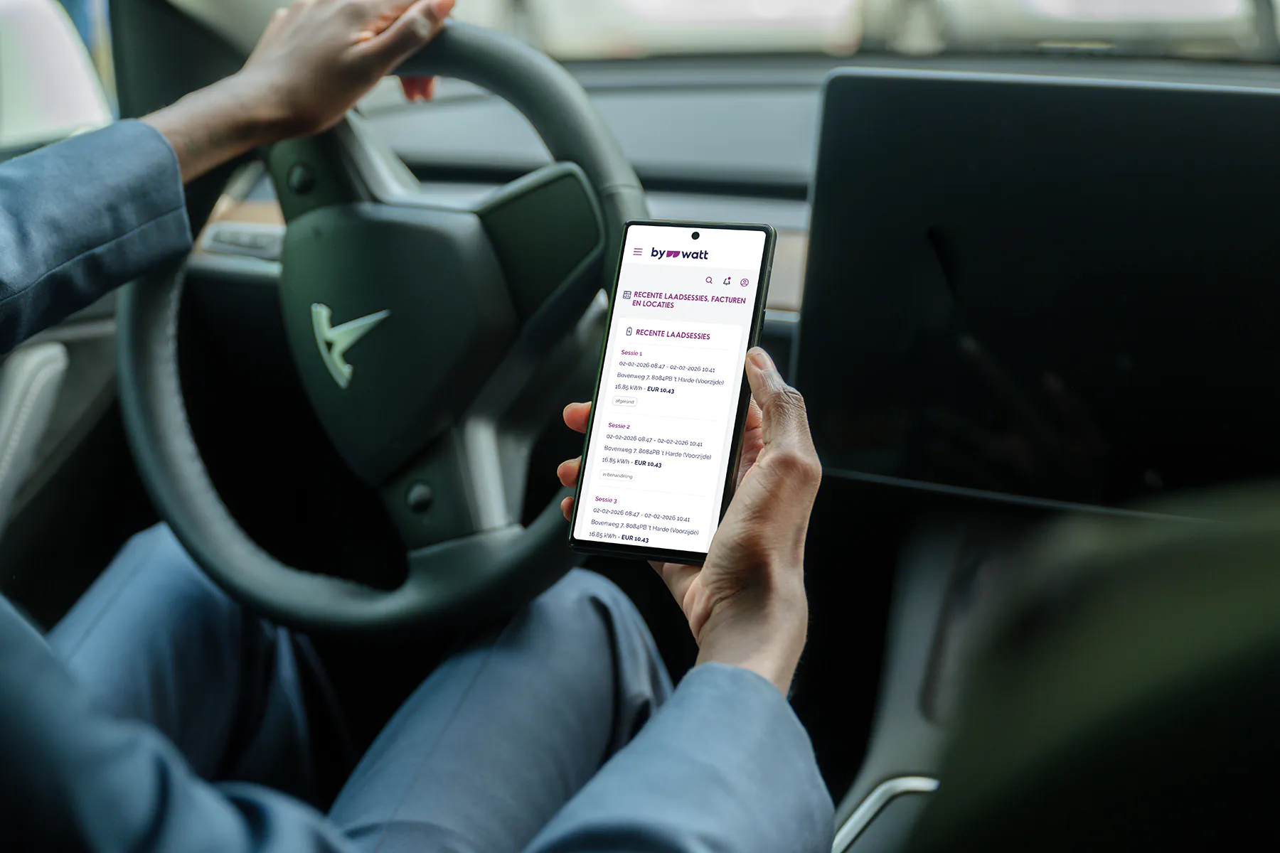 Iemand die in een auto met Tesla-stuur een telefoon vasthoudt met laadstatistieken op het scherm.