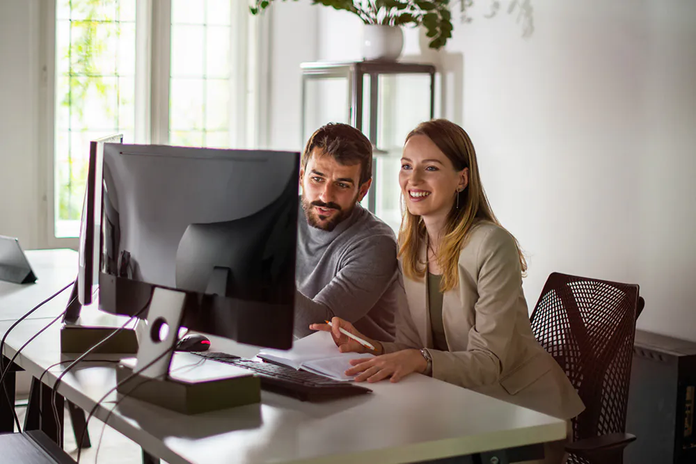 Twee jonge collega's werken samen aan een computer aan een tafel in een lichte kantoorruimte.