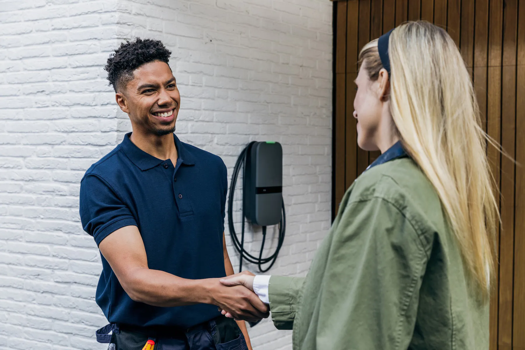 Man in blauw poloshirt schudt hand met vrouw in groene jas voor witte bakstenen muur.