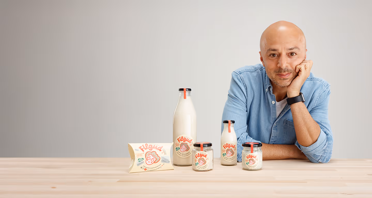Bărbat cu cămașă albastră sprijinit pe masă cu produse lactate Filgud într-o sticlă, borcane și pachet.