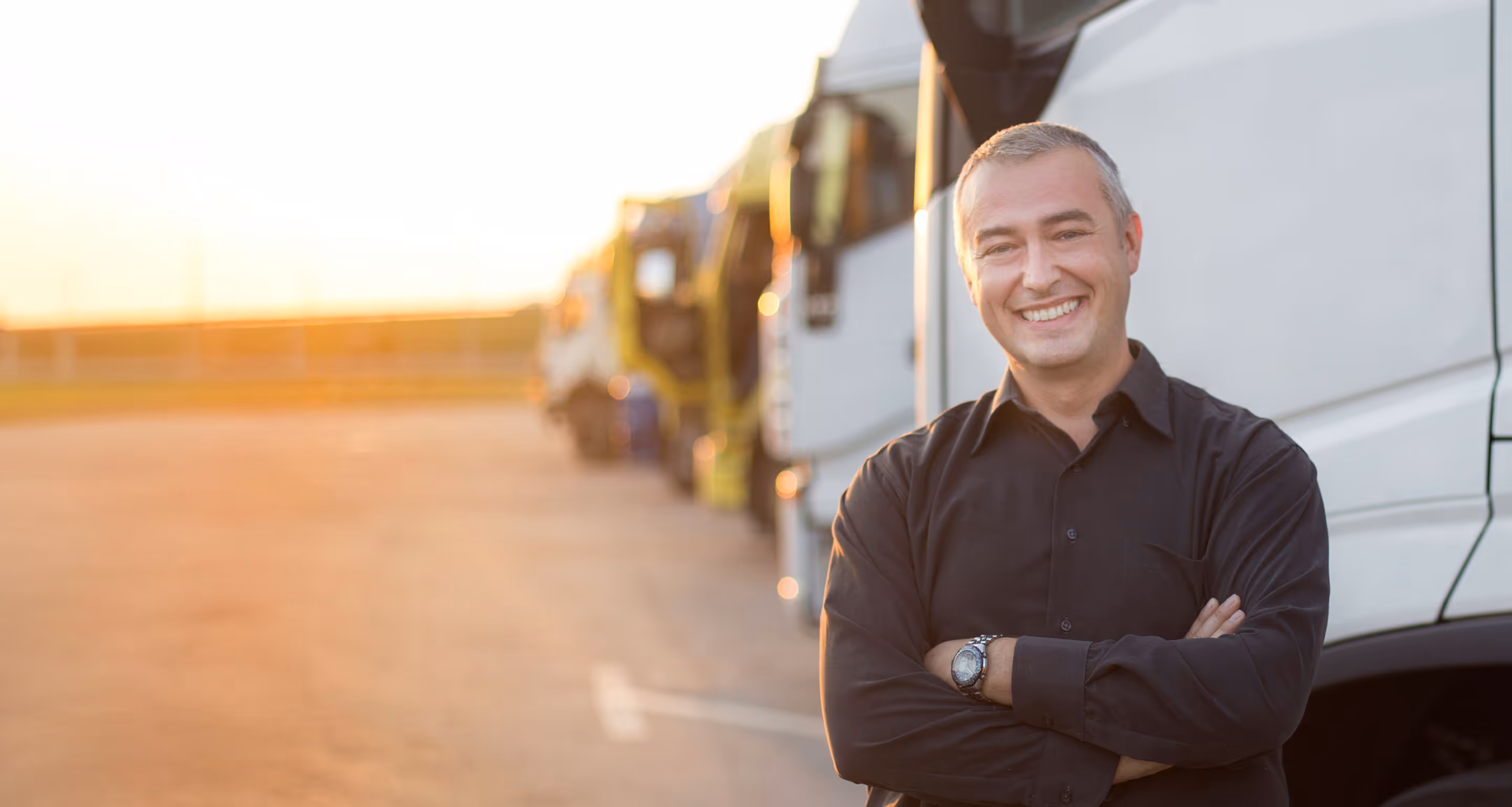 Bărbat zâmbitor în cămașă neagră, cu brațele încrucișate, lângă camioane parcate la apus, pentru factoring în transporturi.