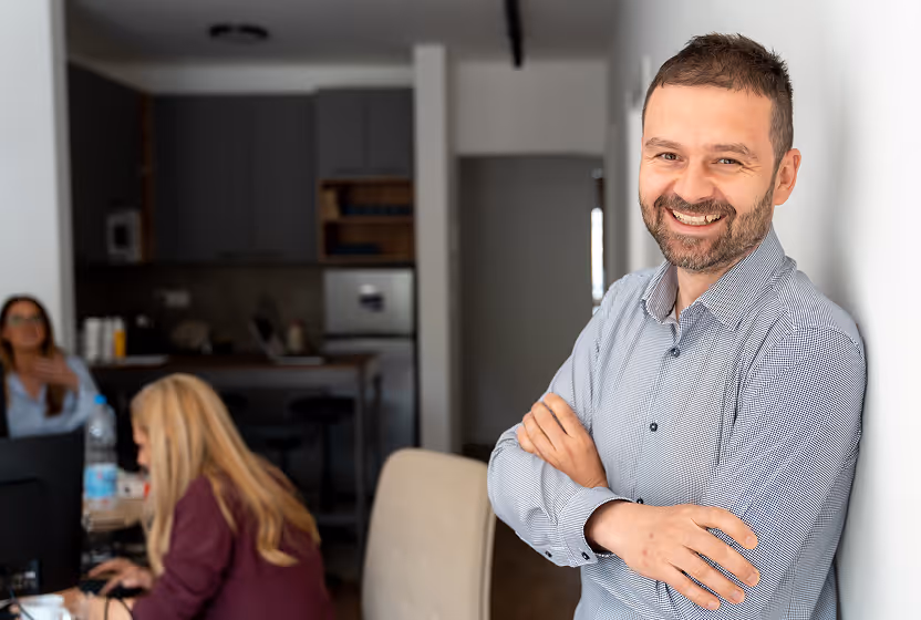 Bărbat zâmbitor cu cămașă, sprijinit de perete, cu două persoane în fundal la Instant Factoring.