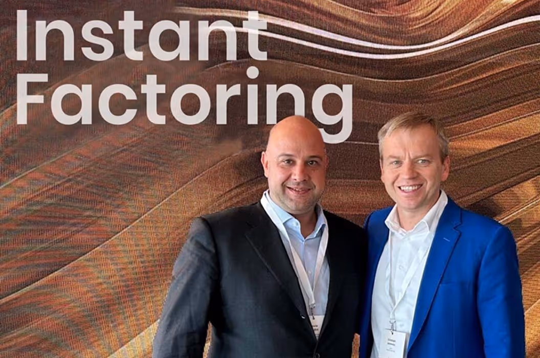 Doi bărbați zâmbind în ținută business, în fața unui fundal Instant Factoring.