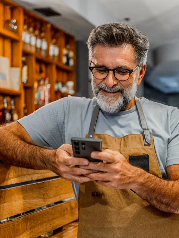 Bărbat cu ochelari și șorț zâmbește folosind telefonul într-un bar, pentru Instant Factoring.