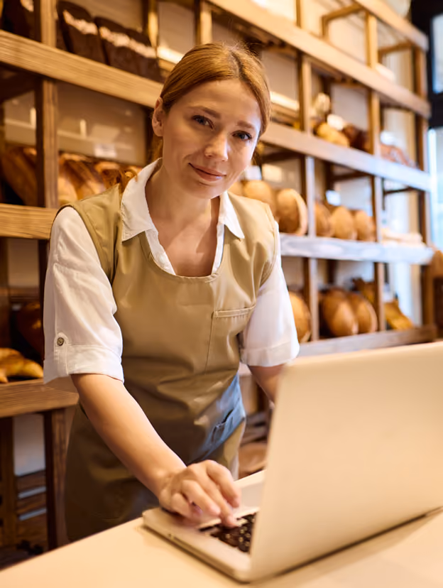 Femeie lucrează pe laptop într-o brutărie, cu pâine pe rafturi, pentru finanțare de afaceri.