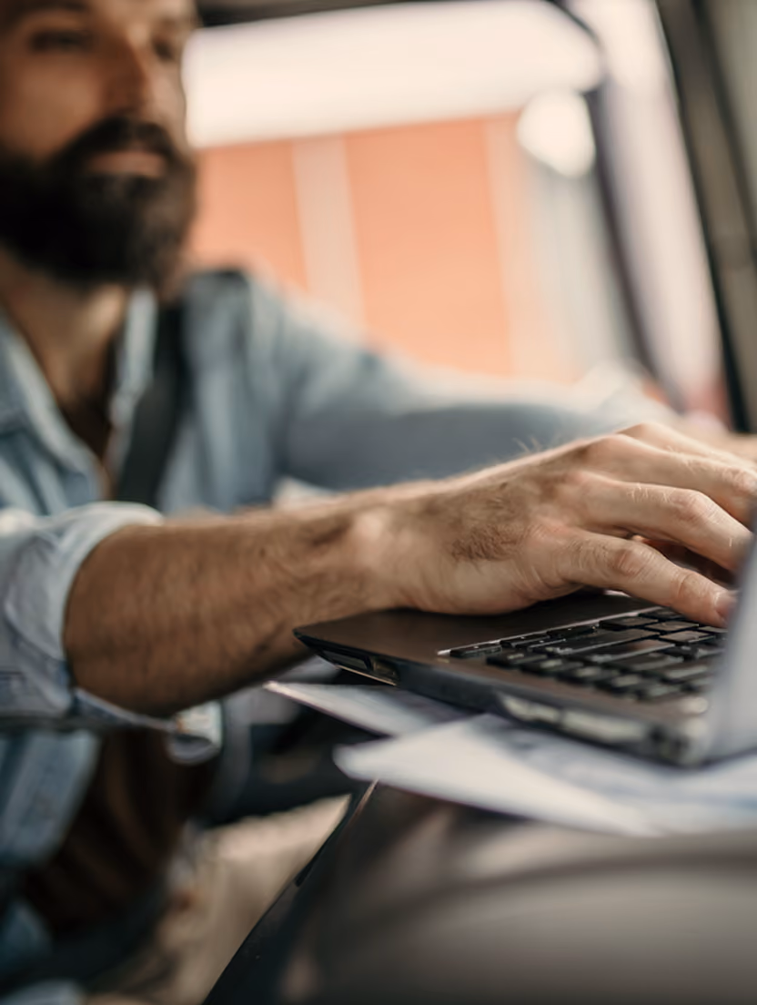 Bărbat cu barbă lucrează la laptop în mașină, verificând opțiuni de finanțare.