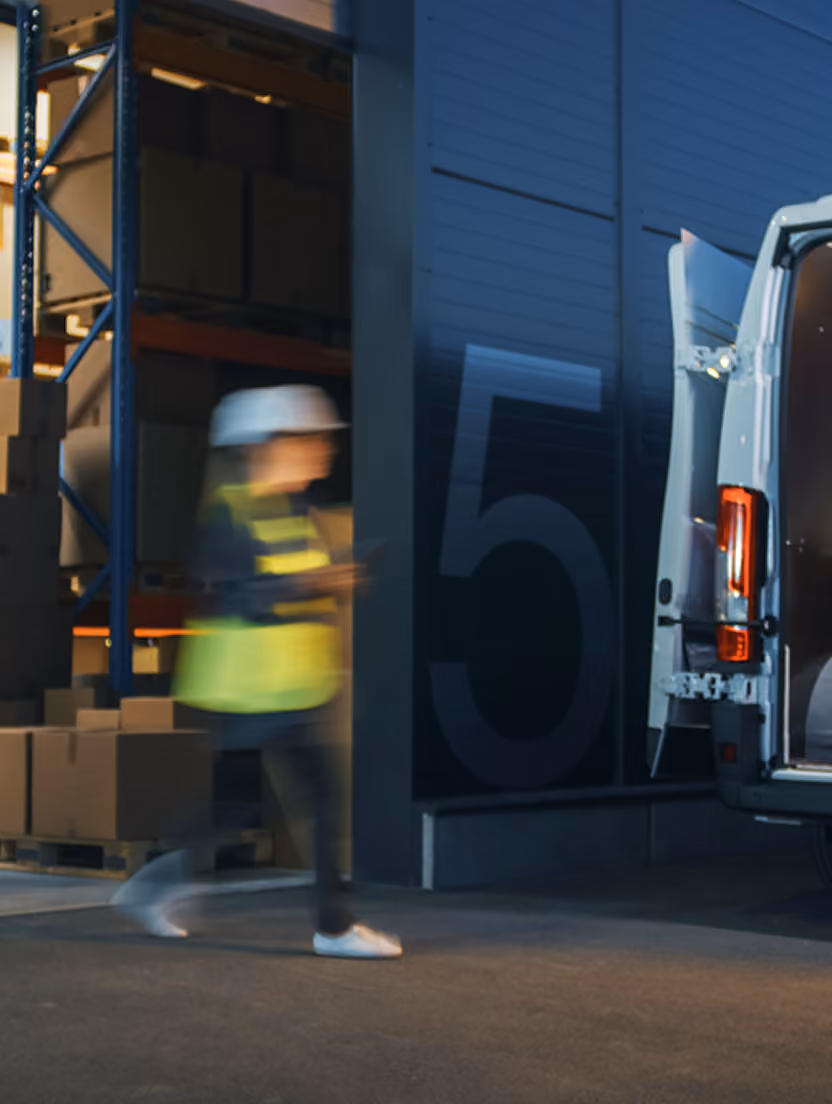 Lucrător cu cască și vestă într-un depozit, lângă o dubă deschisă noaptea, imagine pentru finanțare logistică.