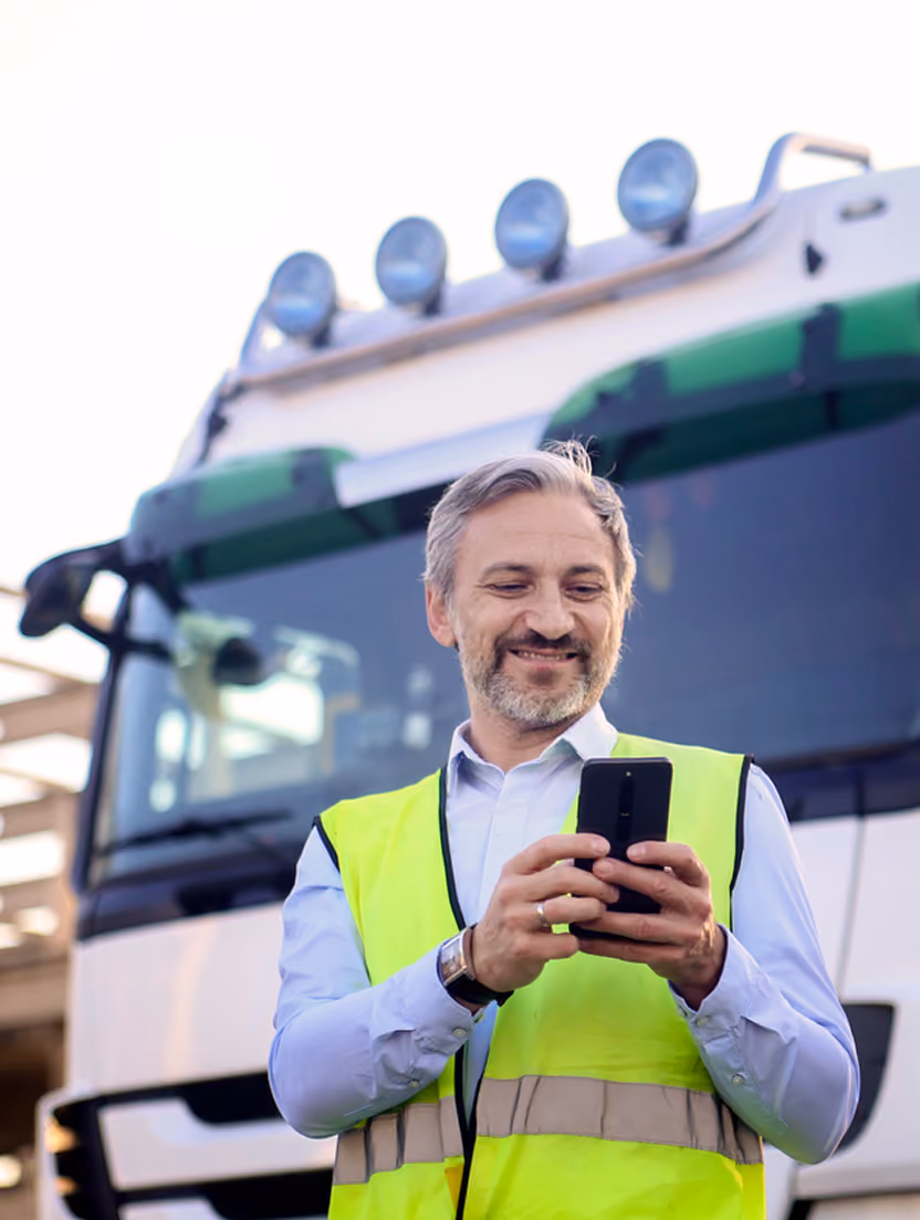 Bărbat zâmbitor cu vestă reflectorizantă folosește telefonul lângă un camion alb, în context de factoring.