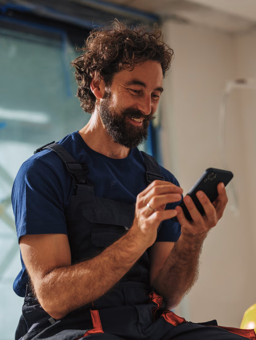Bărbat cu barbă și păr creț, în salopetă, zâmbind cu telefonul mobil pentru Instant Factoring.