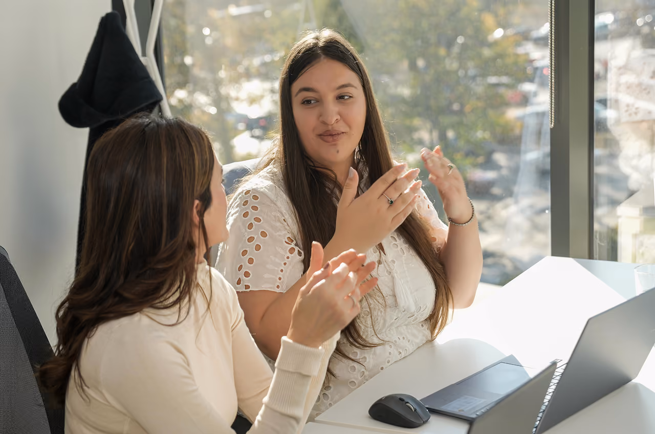 Dos mujeres conversan en una oficina luminosa, gesticulando junto a un portátil en un contexto de factoring