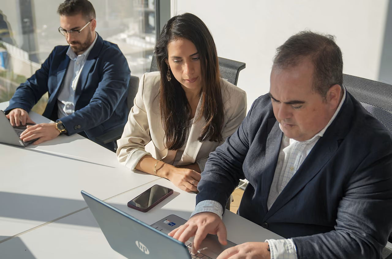Tres personas en una reunión de factoring, usando portátiles en una mesa blanca en una oficina luminosa.
