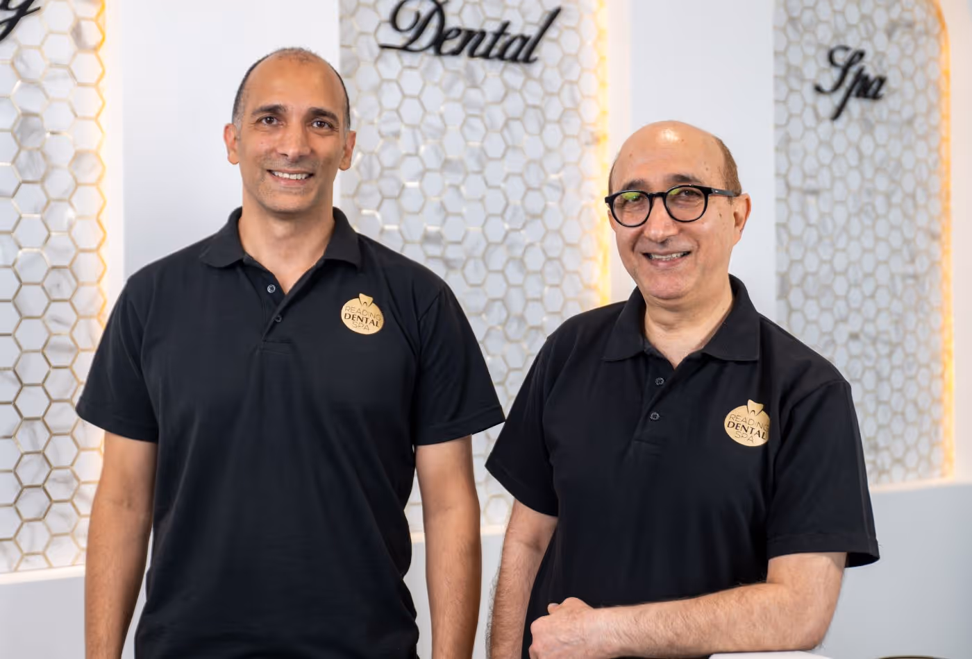 Two men in black polo shirts with Reading Dental Spa logos smiling in front of honeycomb wall panels.