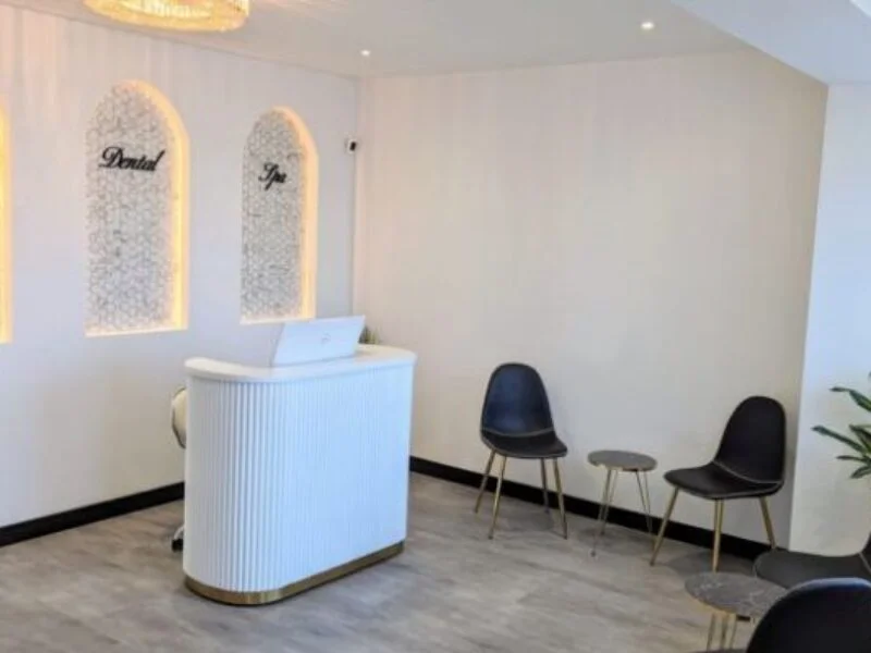 Bright reception area with white curved desk, two black chairs, small tables, and wall signs 'Dental' and 'Spa'.