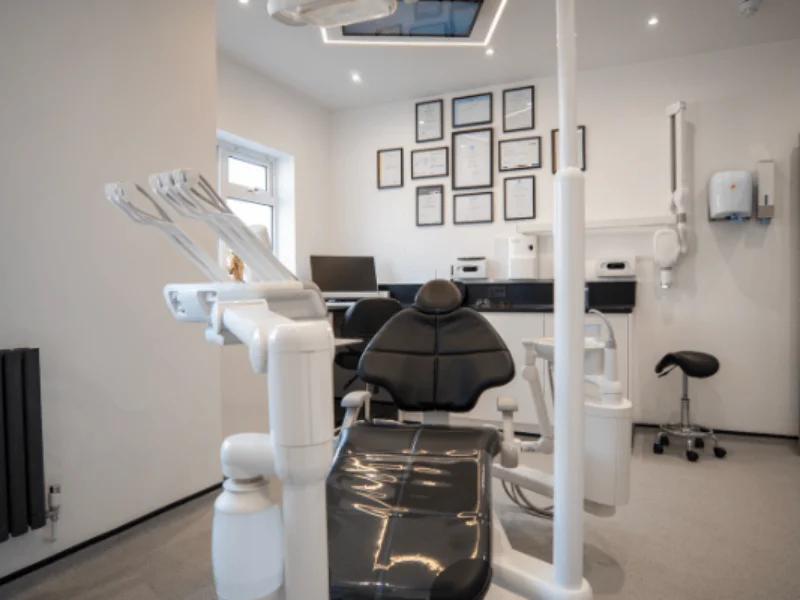 Modern dental clinic with a black dental chair, equipment, computer desk, and framed certificates on the wall.