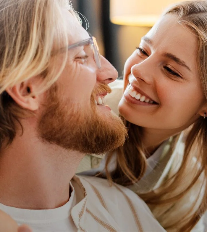 Smiling woman looking affectionately at a bearded man wearing glasses indoors.
