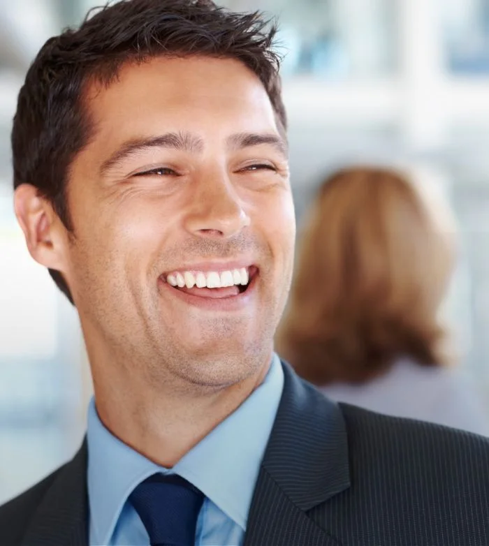 Smiling man in business suit and blue shirt with blurred person in background.