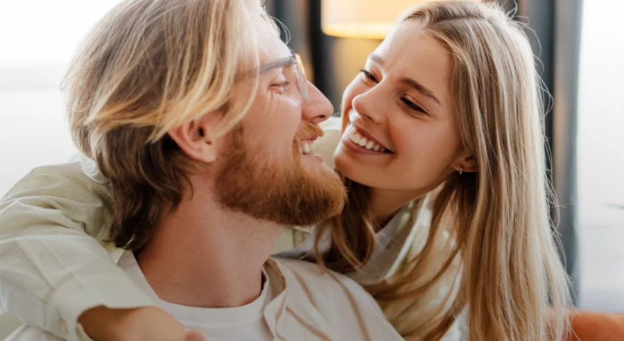 Smiling woman hugging and leaning close to a man with glasses and a beard indoors.