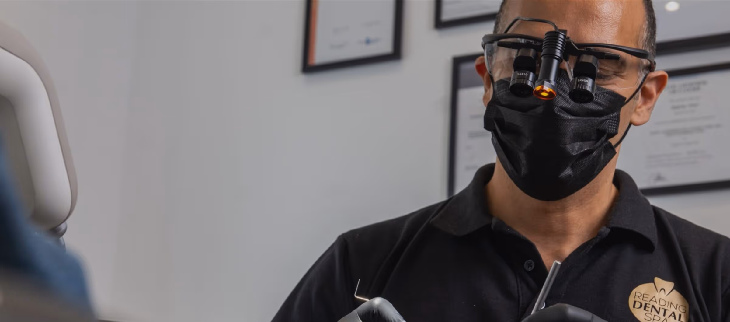 Dentist wearing magnifying glasses, black mask, and gloves working with dental tools in clinic.