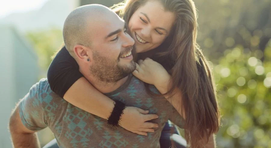 Smiling woman hugging a man from behind outdoors in sunlight with blurred green background.
