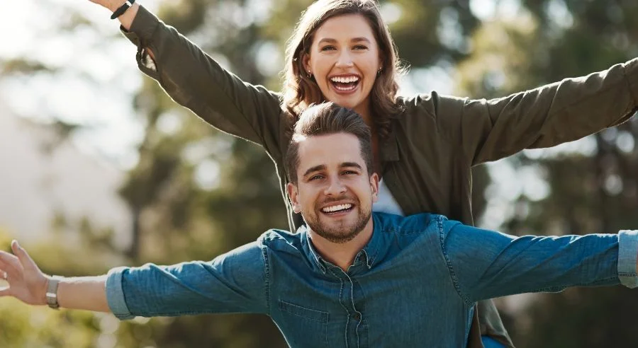 Smiling man and woman outdoors with arms outstretched, enjoying a sunny day together.